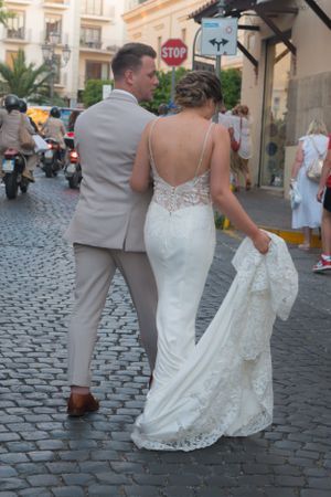 A newlywed bride and groom walking through a busy Italy street. The groom is looking at the bride and she is holding her dress up.