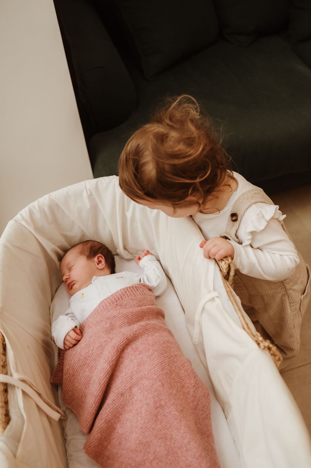 A sleeping newborn baby wrapped in a pink blanket lies in a cream-colored bassinet while being watched over.