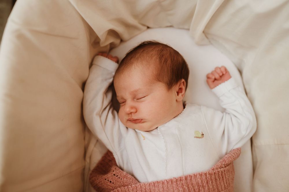 Peaceful sleeping newborn baby wearing white top and pink blanket rests on cream colored bedding.