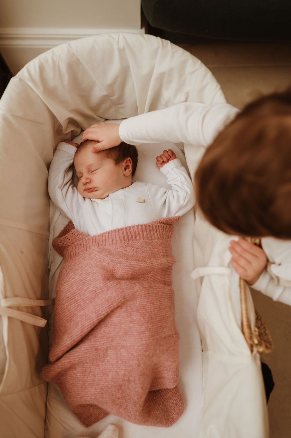 A sleeping newborn baby wrapped in a pink blanket rests peacefully in a white bassinet.