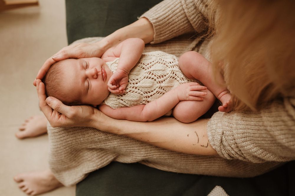 A sleeping newborn wearing a knit romper rests peacefully in gentle hands on a cozy blanket.
