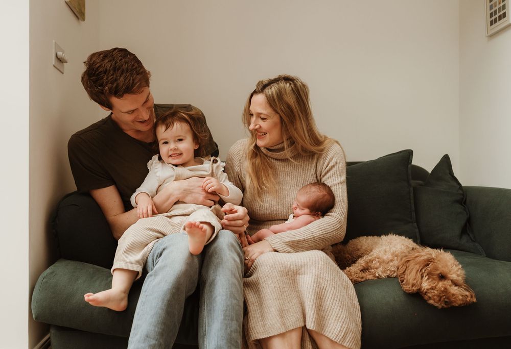 A family sits together on a dark green couch with their dog while holding a newborn baby and toddler.