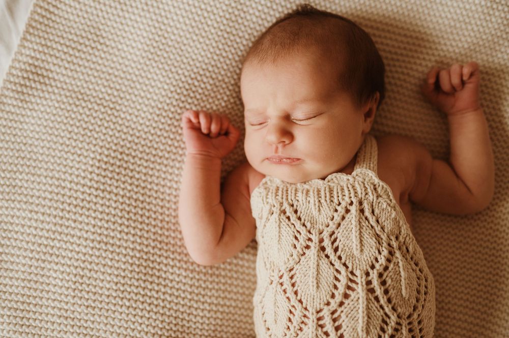 Sleeping infant wearing cream knit dress lies peacefully on textured beige blanket with arms raised.