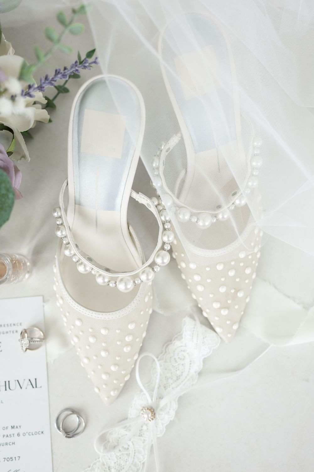 White bridal shoes adorned with pearl details displayed alongside wedding stationery and floral elements.