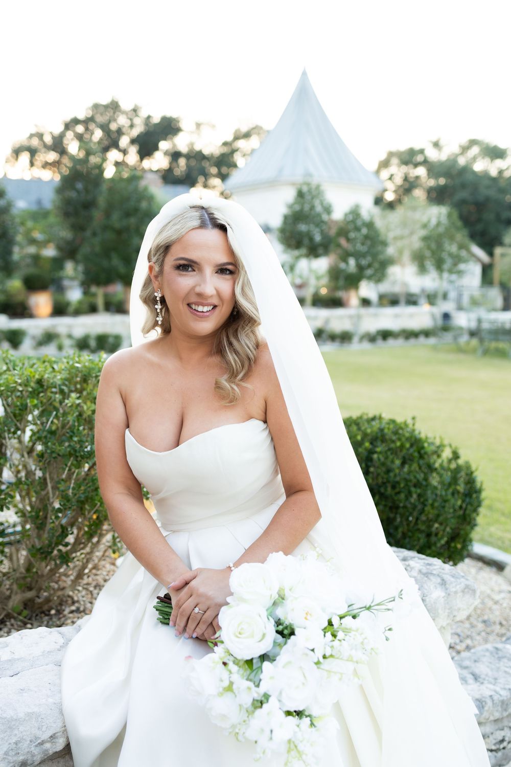 Beautiful bride in strapless white gown holds elegant floral bouquet at outdoor garden venue with chapel spire in background.