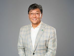 A person wearing glasses and a plaid blazer smiles warmly against a gray background.