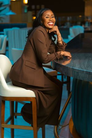 Person in a brown suit sits casually in a white chair at a restaurant, showing a joyful expression.
