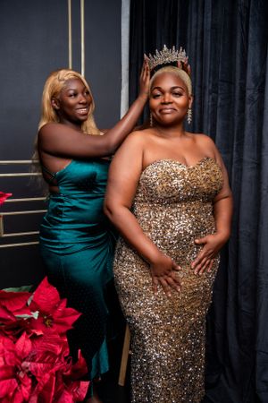 Two women in evening gowns pose together with tiara and poinsettias.