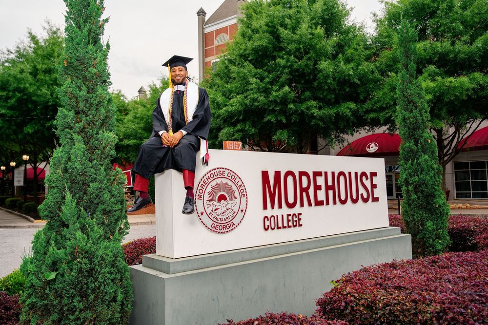 Graduate in cap and gown sitting on Morehouse College sign, surrounded by lush green trees.