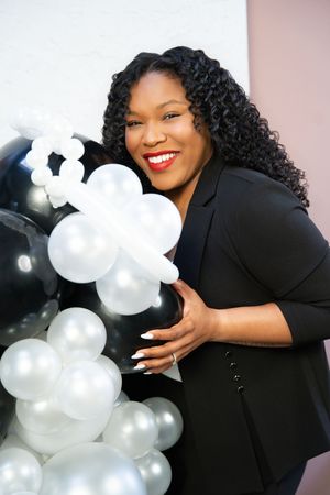 A person in a black dress poses playfully with white and silver balloon arrangements against a pink backdrop.