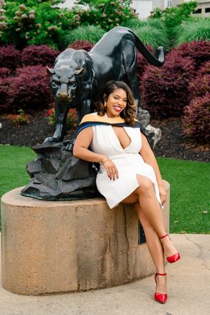 Woman in white dress and red heels poses by a black panther statue outdoors at a graduation celebration.