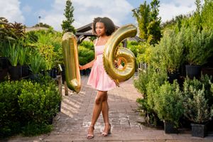 Young girl in pink dress celebrates sweet 16 with gold number balloons in garden setting.