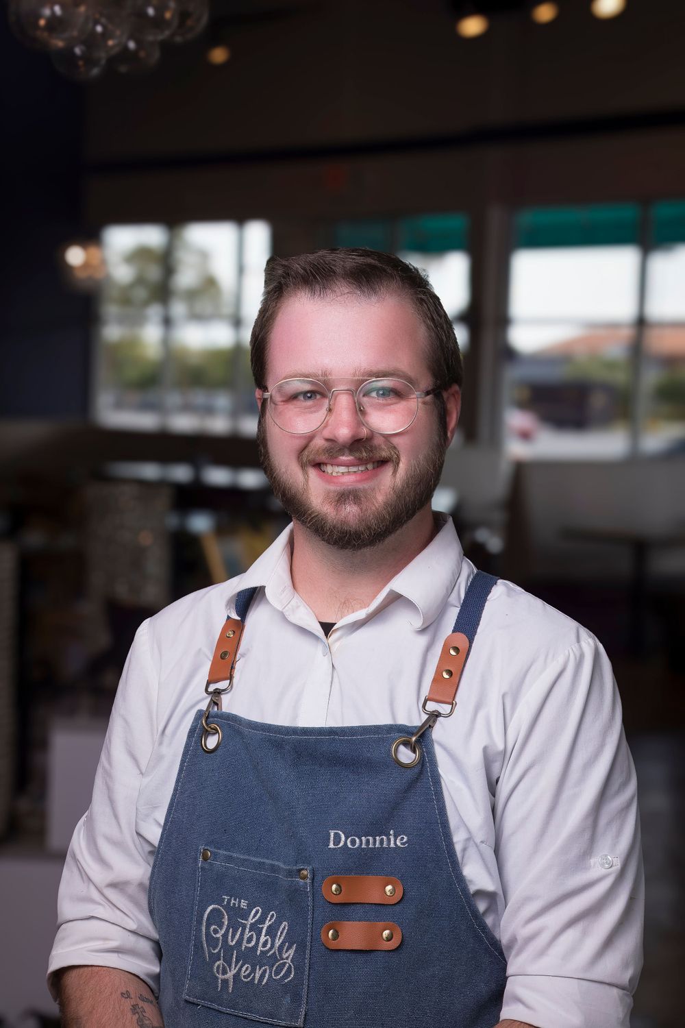 Restaurant worker wearing denim apron with leather straps and white shirt in dining room setting.