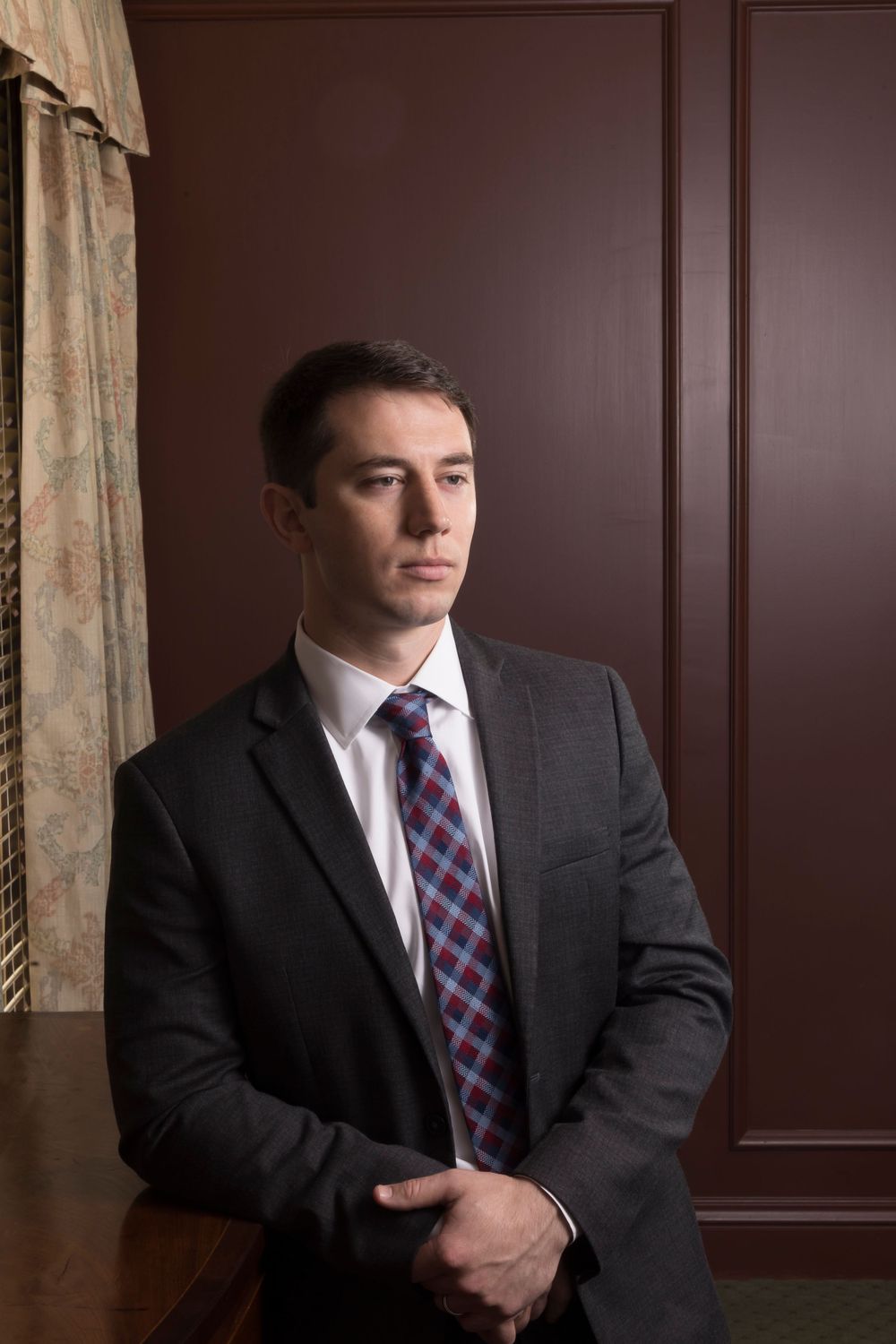 Professional portrait of a person in dark suit with plaid tie against burgundy wall.