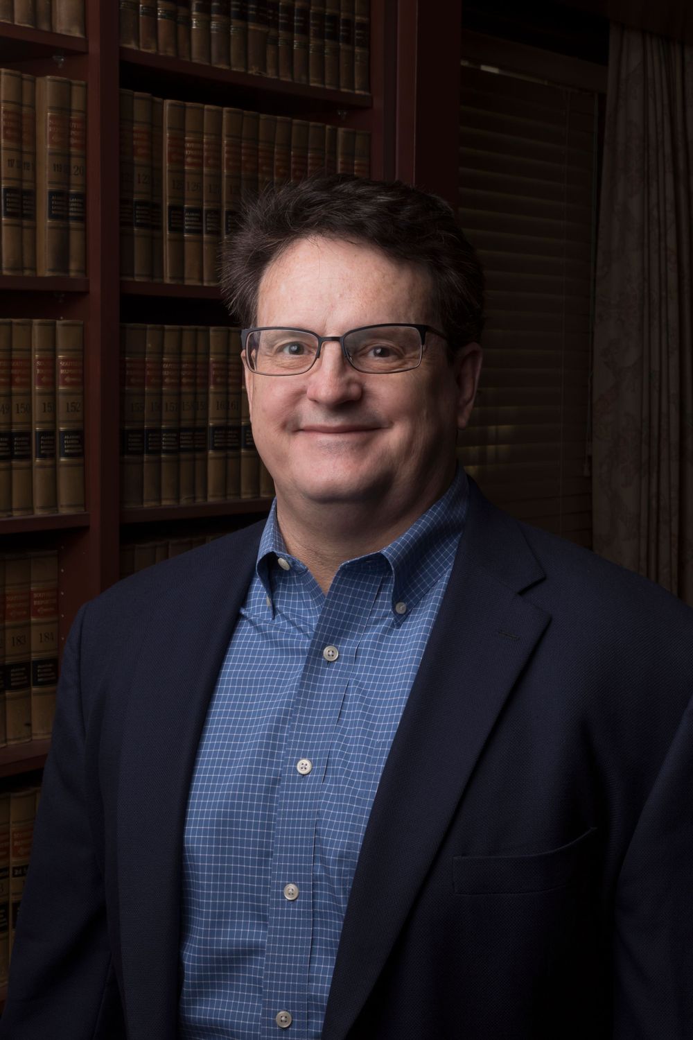 Professional portrait of a person in blue button-down and blazer with glasses in library setting.