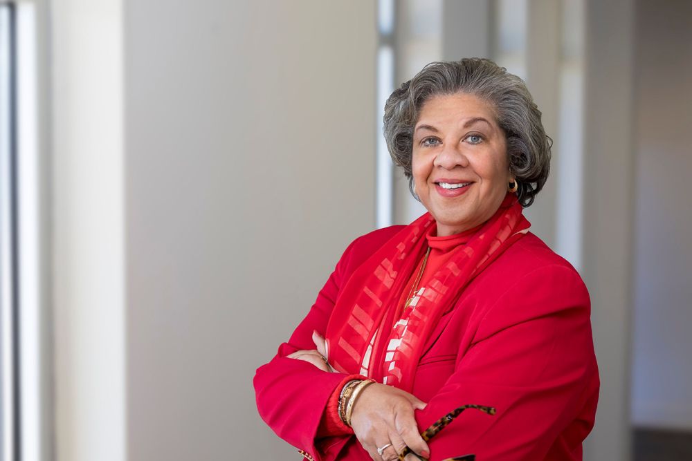 Portrait of a person in a bright red jacket standing by a window with a cheerful expression.