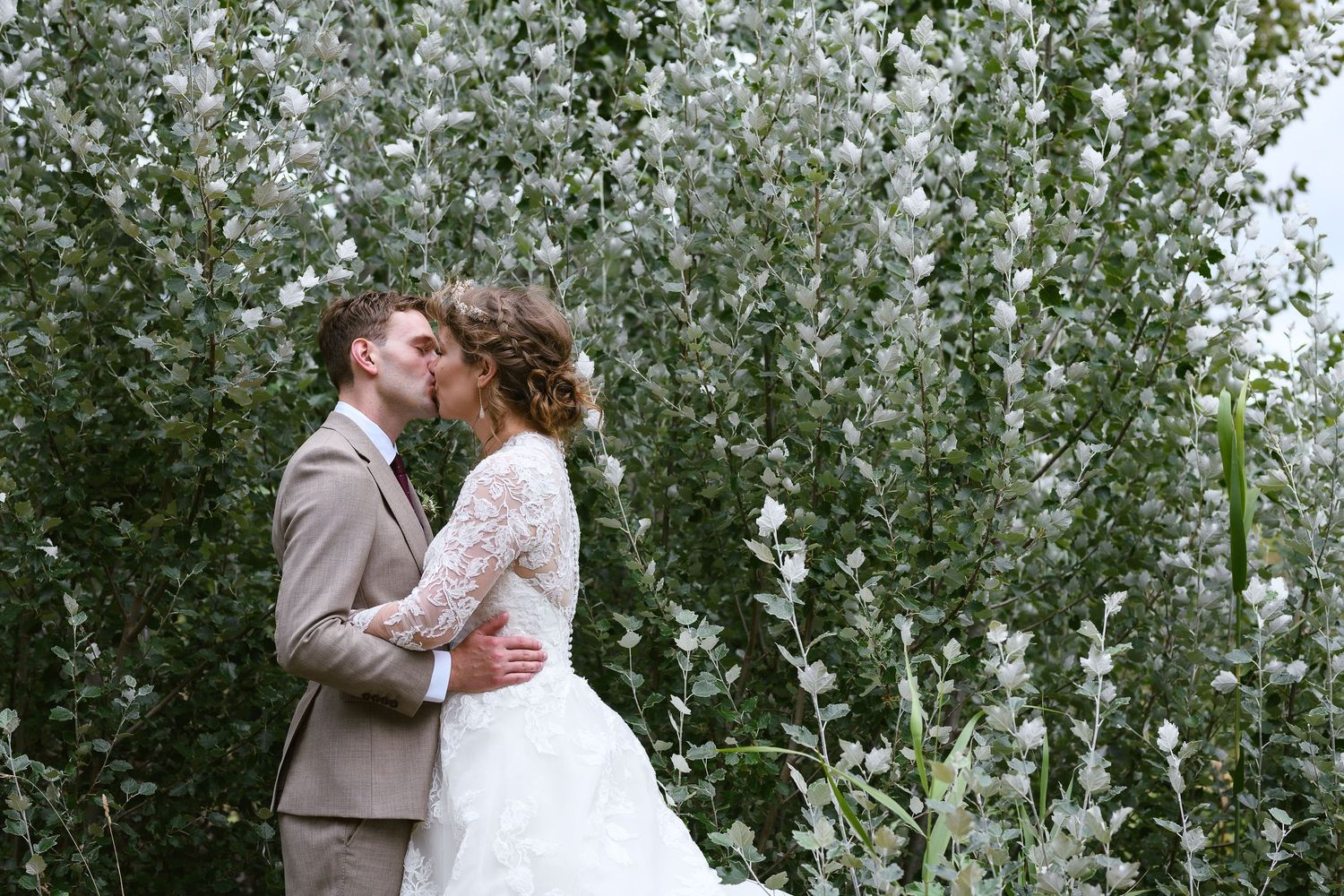 Romantic wedding couple embraces in front of white flowering trees during outdoor ceremony.