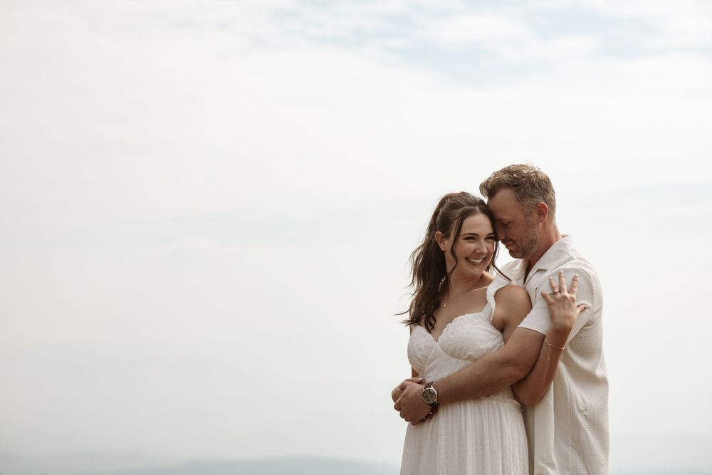 A romantic couple in white clothing embraces tenderly during a cloudy outdoor photo session.