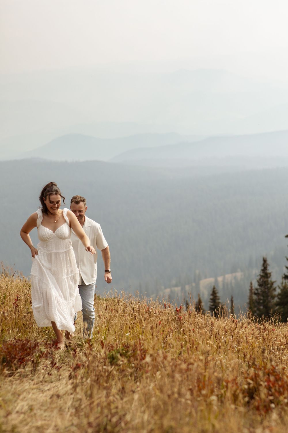 A couple walks together through a golden mountain meadow with misty mountain ranges visible in the distance.