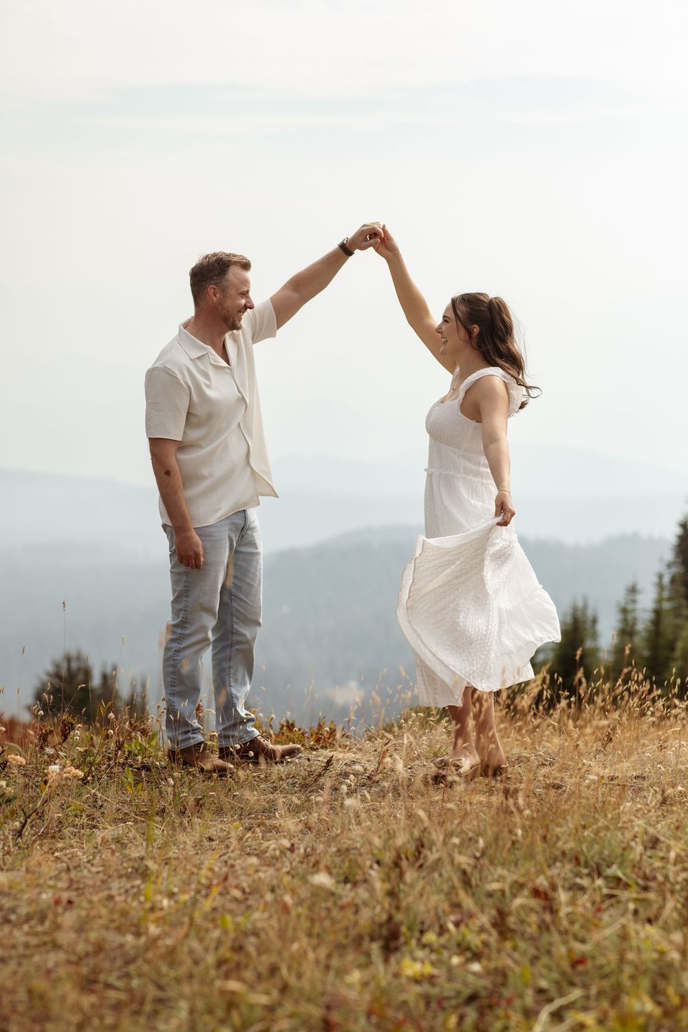 A couple in white outfits dance together on a hilltop meadow with mountain views in the background.