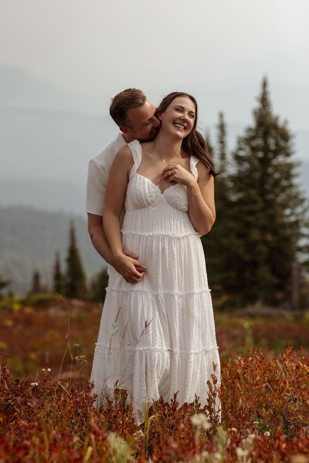 A couple in white dresses embraces in a field of red autumn foliage with evergreen trees and mountains in the background.