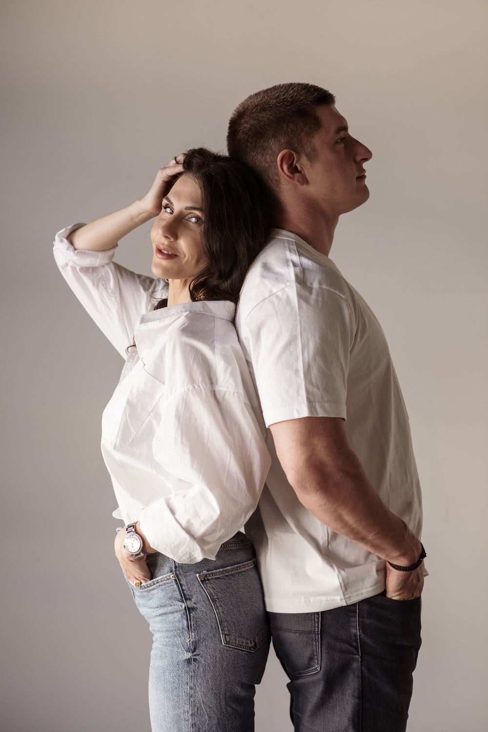 Artistic fashion photo series showing couple in white shirts and jeans posing back to back against neutral wall.