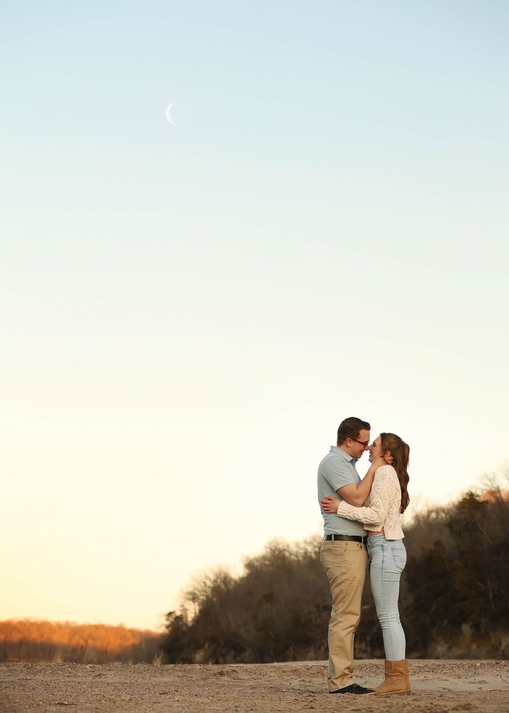 A couple stands together against a sunset sky on an open beach landscape.