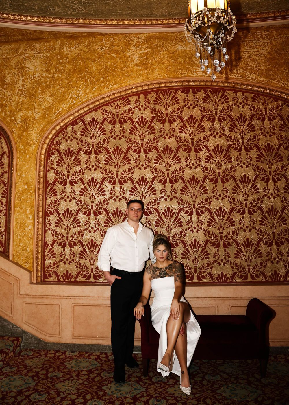 A couple posing against an ornate damask wallpaper in formal attire.