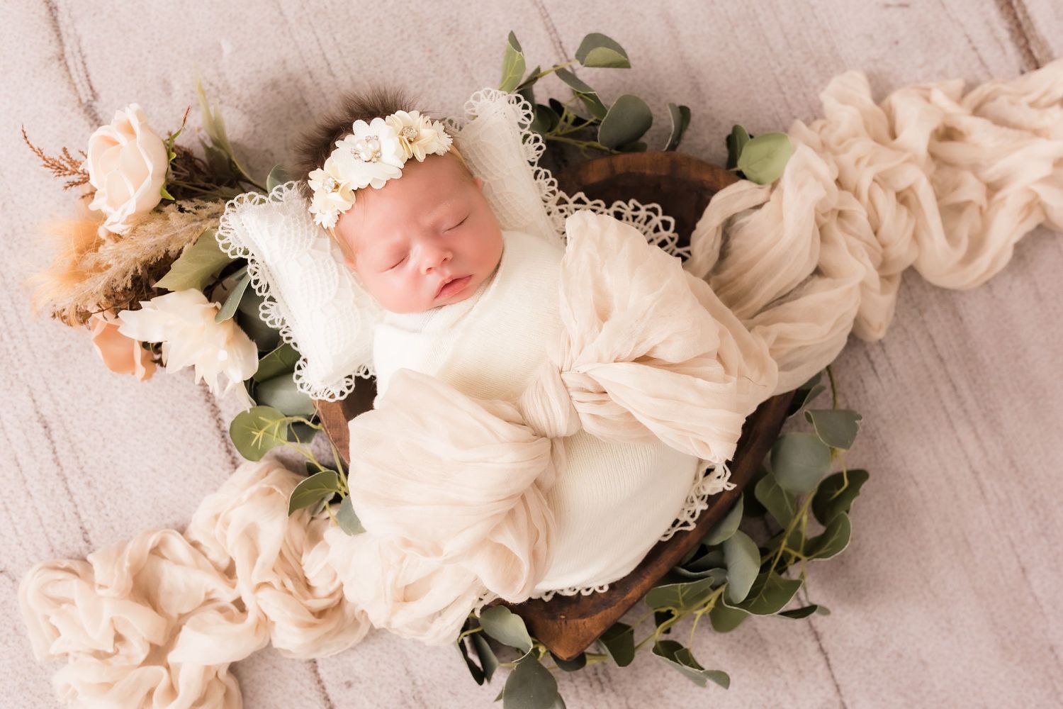 Sleeping newborn baby swaddled in cream wrap with floral headband in rustic wooden bowl with greenery.