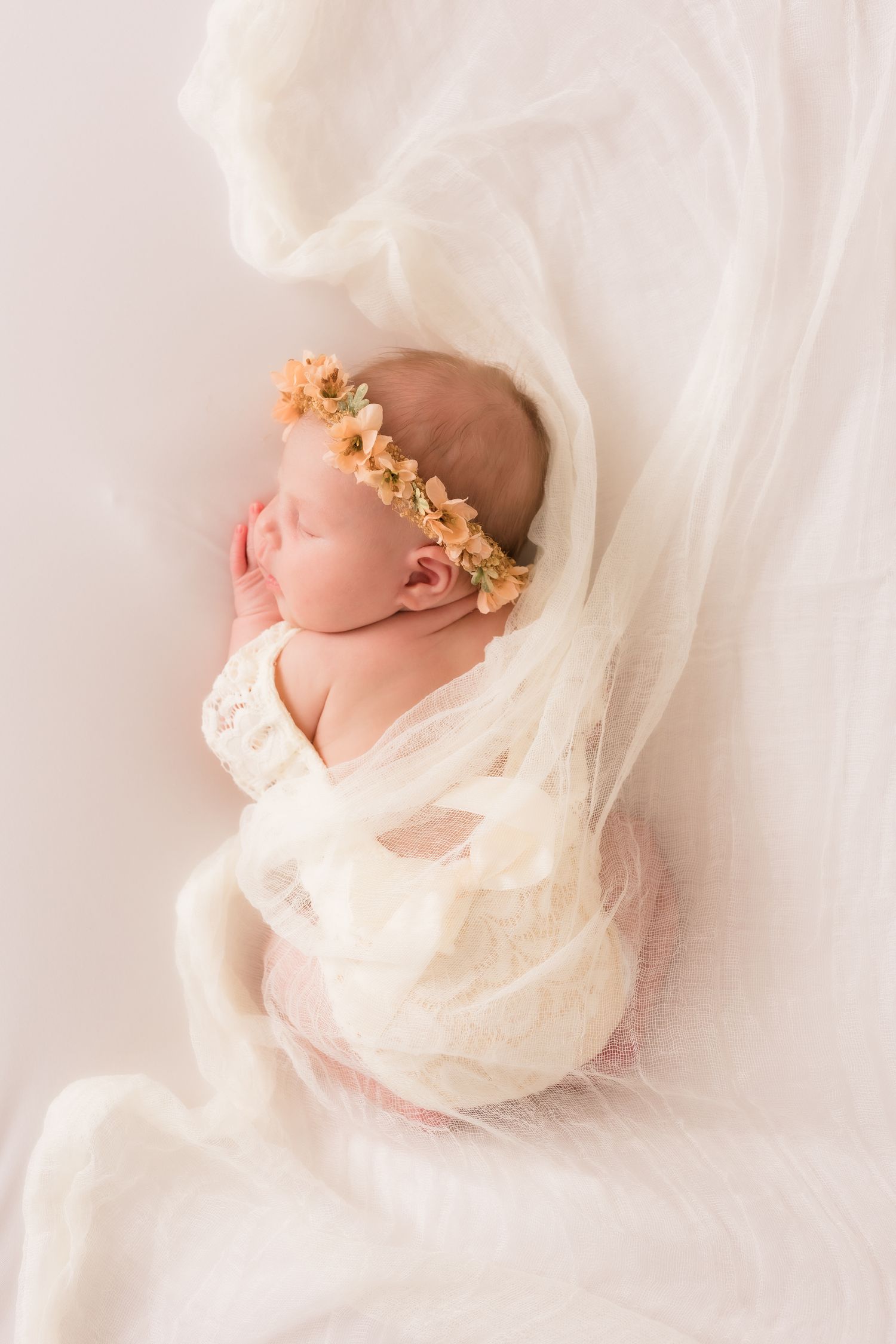 Newborn baby girl sleeping wrapped in white lace swaddle with peach floral crown on soft backdrop.