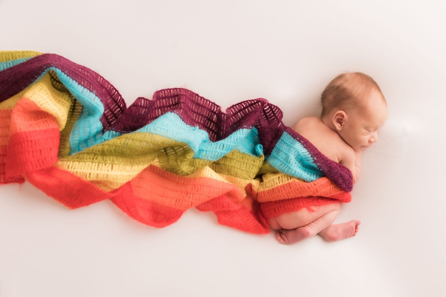 Peaceful newborn baby sleeping wrapped in a vibrant rainbow striped knit blanket on white background.