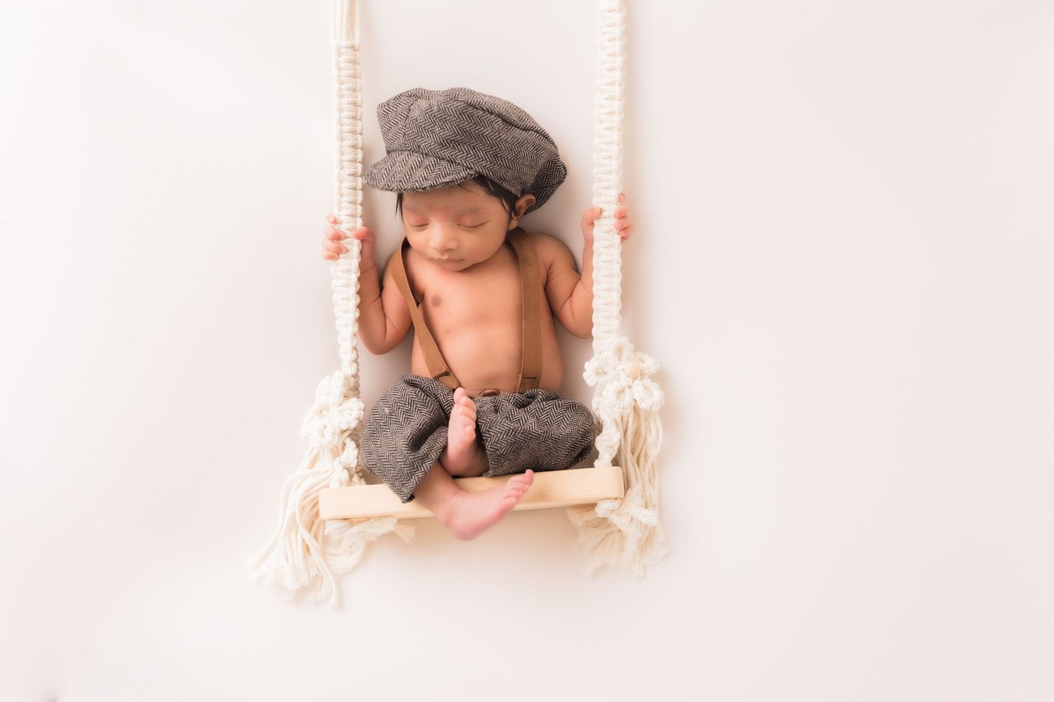 Sleeping newborn baby wearing gray newsboy cap and pants posed on a macrame swing prop.