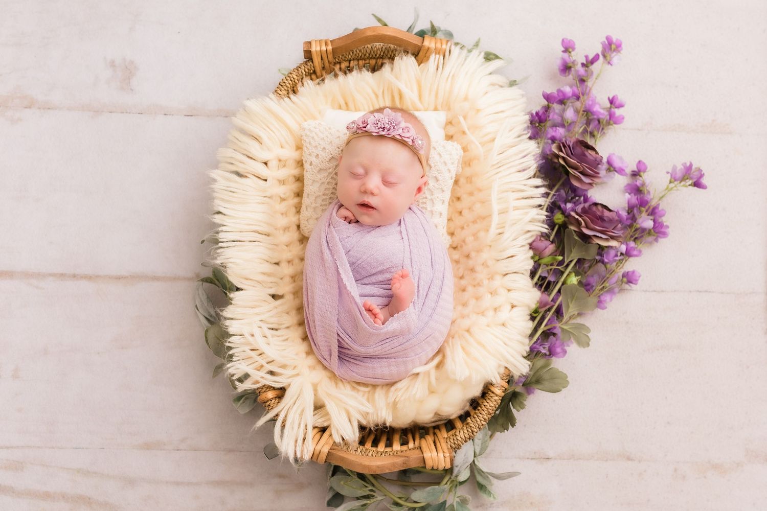 Sleeping newborn baby girl wrapped in lavender swaddle in wicker basket with purple flowers.