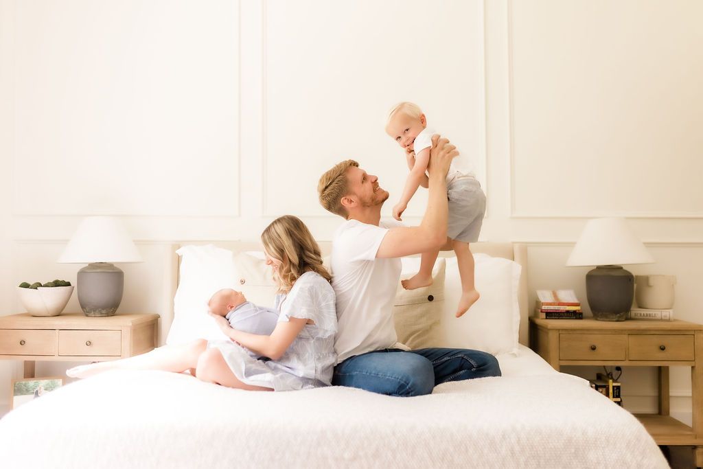 A father lifts a baby joyfully on a white bed while a mother holds another child in a bright, cozy bedroom.
