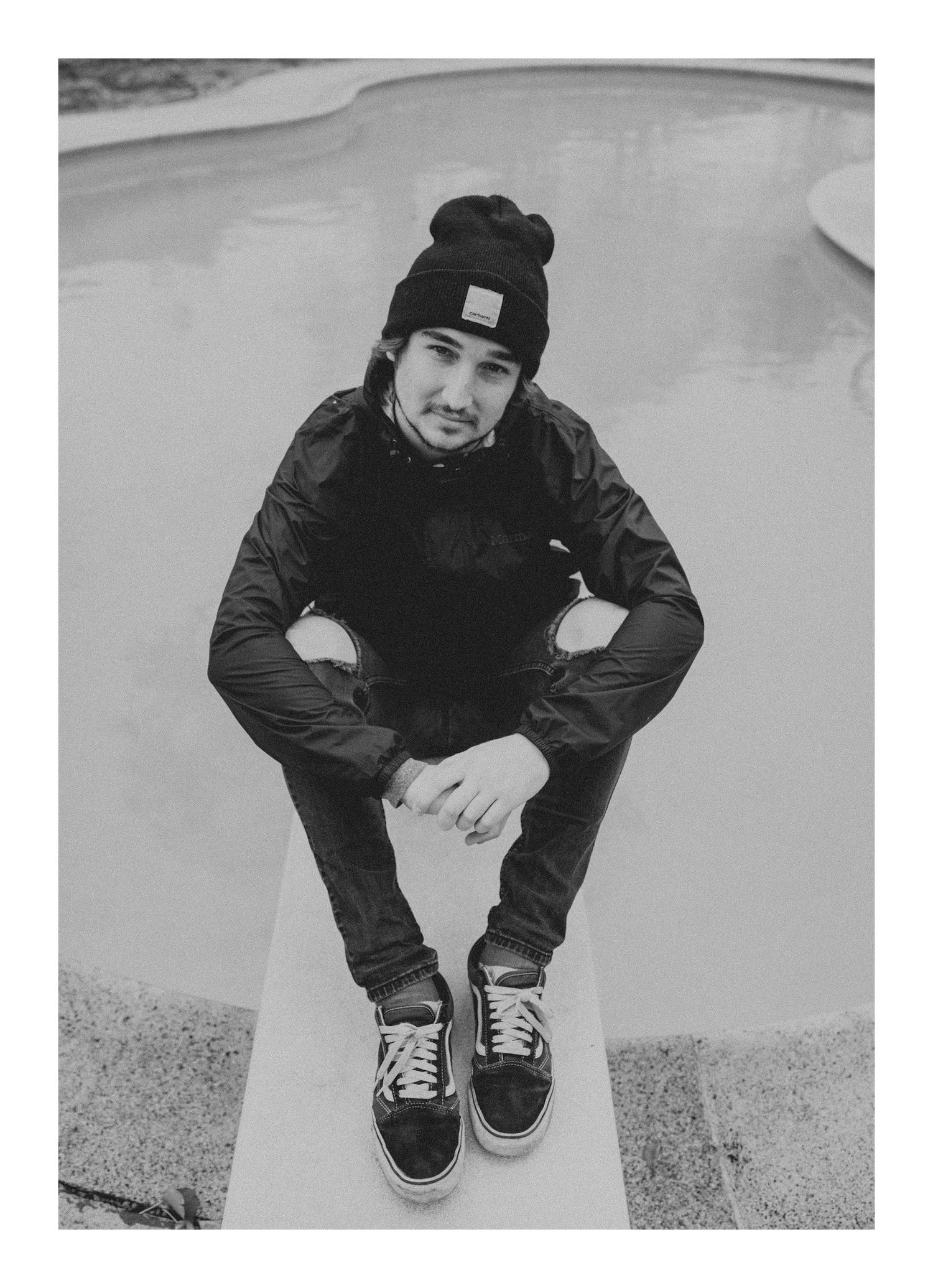 Person wearing black beanie and jacket sitting in an empty concrete skate park.