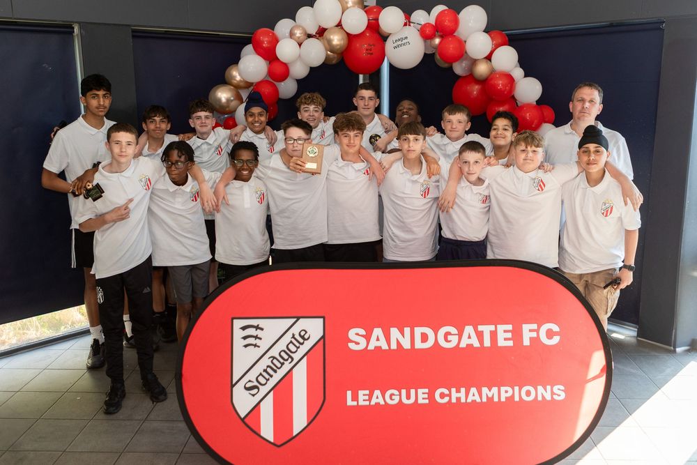 Sandgate FC youth league champions team photo with boys in white shirts celebrating in front of branded signage.