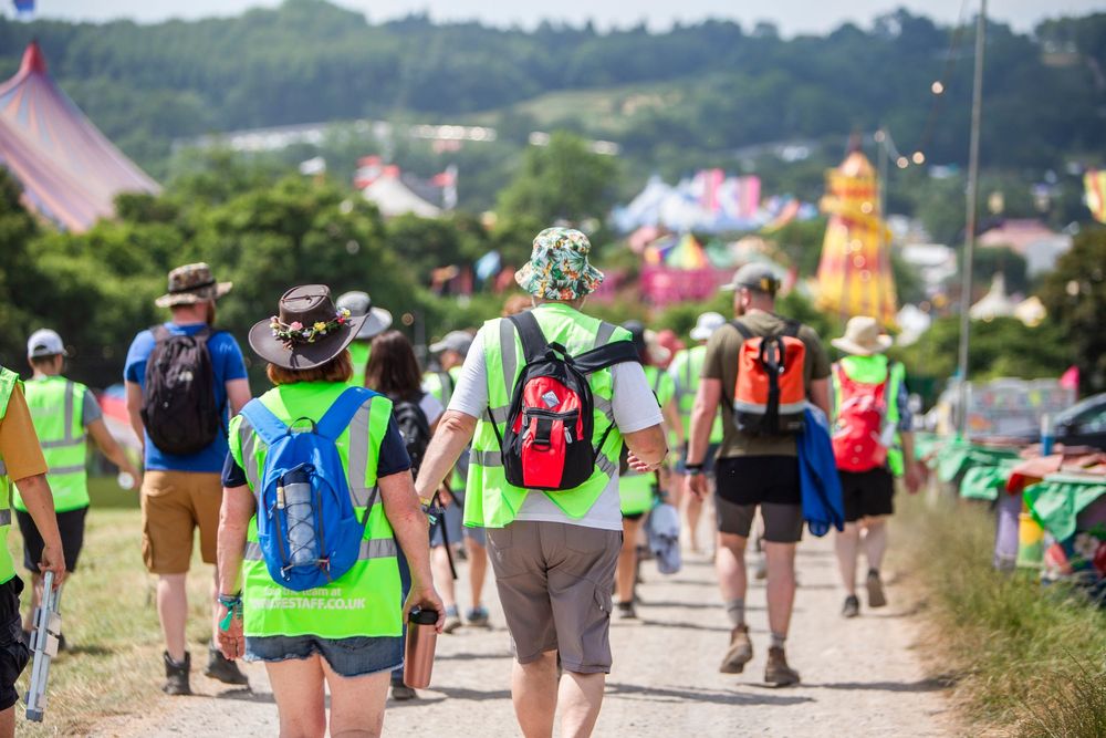 Festival volunteers in hi-vis vests walk along a dusty path toward colorful tents and attractions on a sunny day.