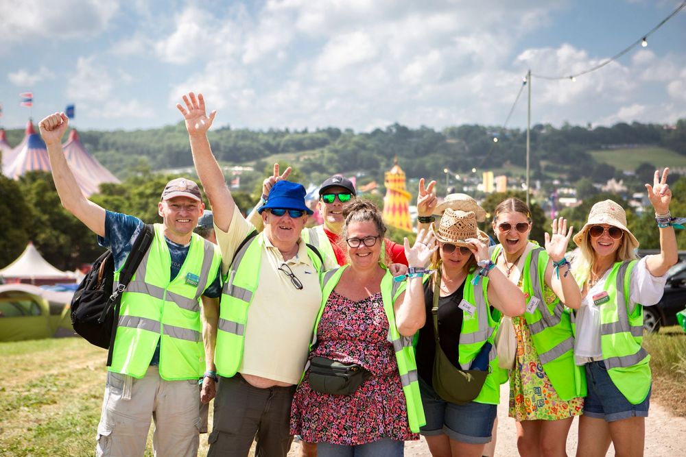 Group of smiling festival volunteers in high-vis vests waving at camera outdoors on a sunny day.