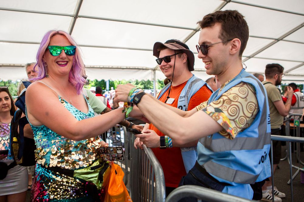 Festival staff scanning wristbands at entrance gate, with colorful attendees in sequin outfit and sunglasses.