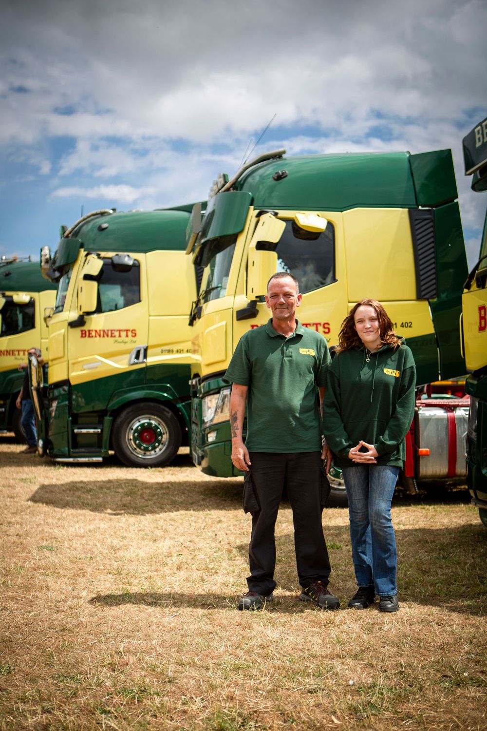 Two Bennetts staff members stand proudly in front of a fleet of green and yellow Bennetts trucks outdoors.