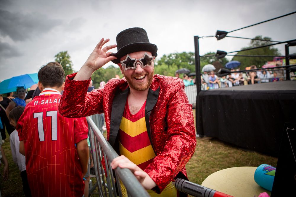 Man in red sequin jacket, star sunglasses, and black top hat poses at outdoor festival near wrestling ring.