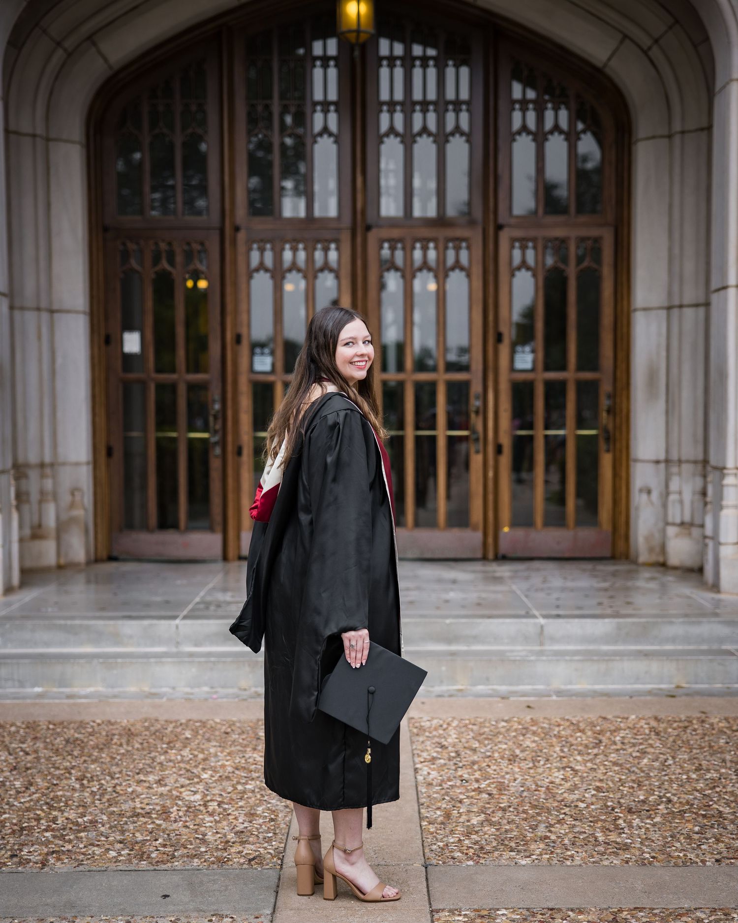A smiling graduate in black cap and gown poses in front of ornate wooden doors at a university building.