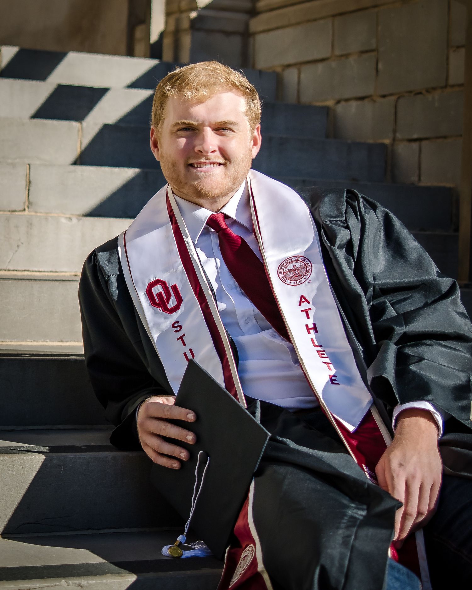 Graduate in black gown and red stole sits for professional graduation portraits in a stone archway.