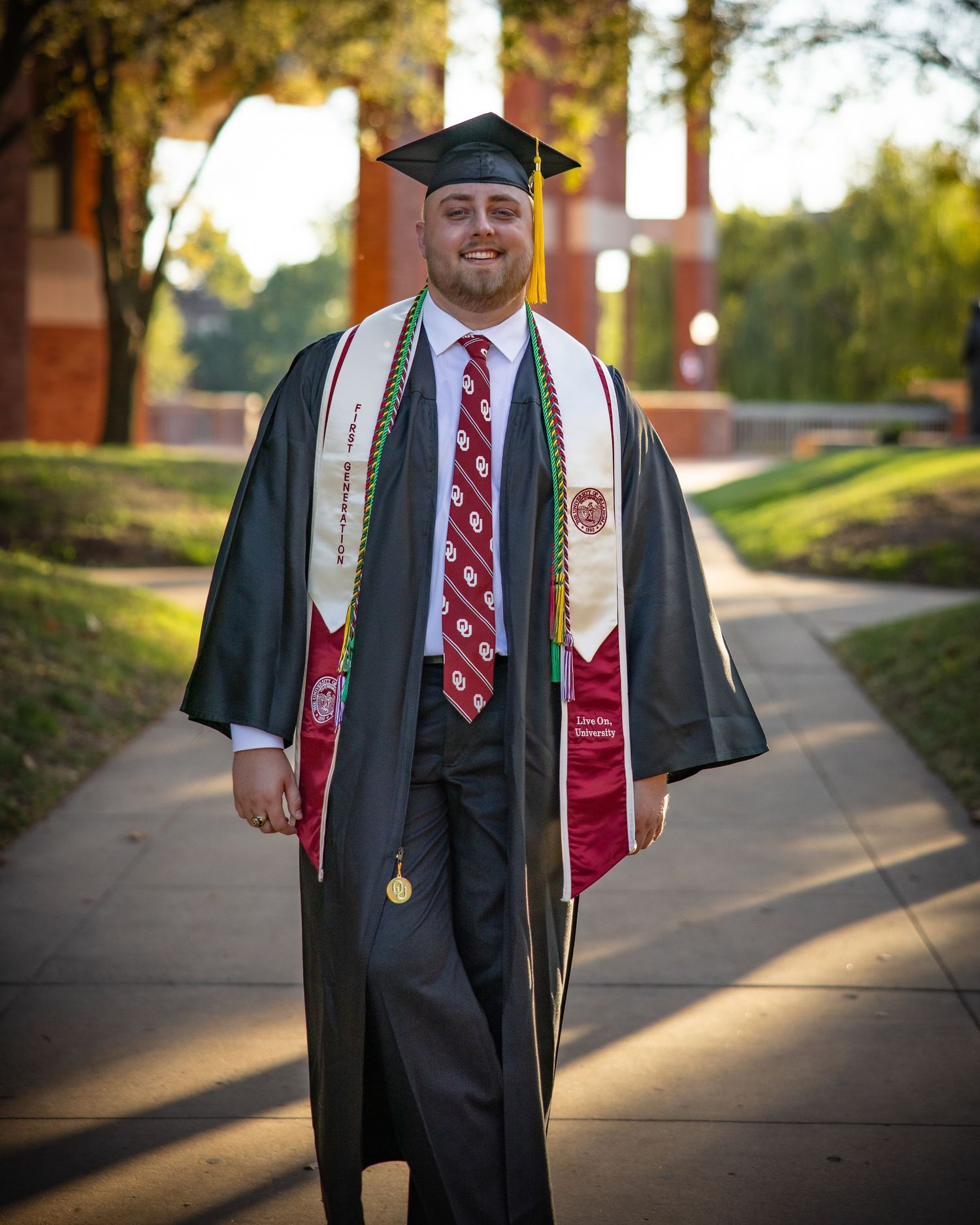 Graduate in black cap and gown with red and white stole stands on sunlit campus walkway during graduation photoshoot.