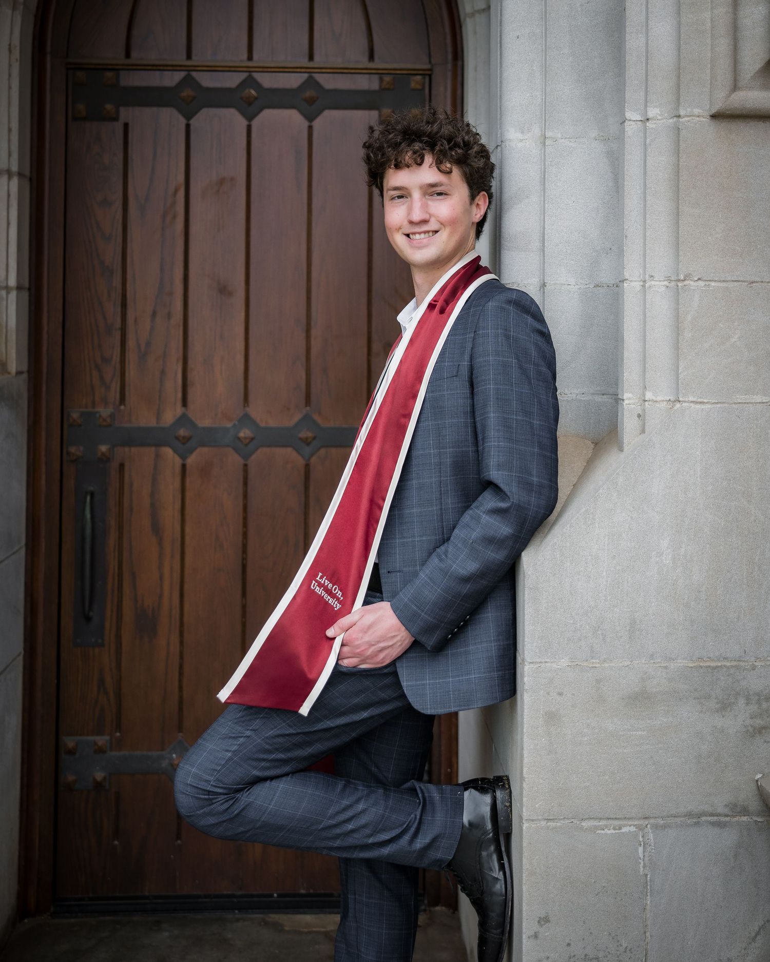 Young man in gray suit with red graduation stole leans against stone wall by wooden door for senior portrait.