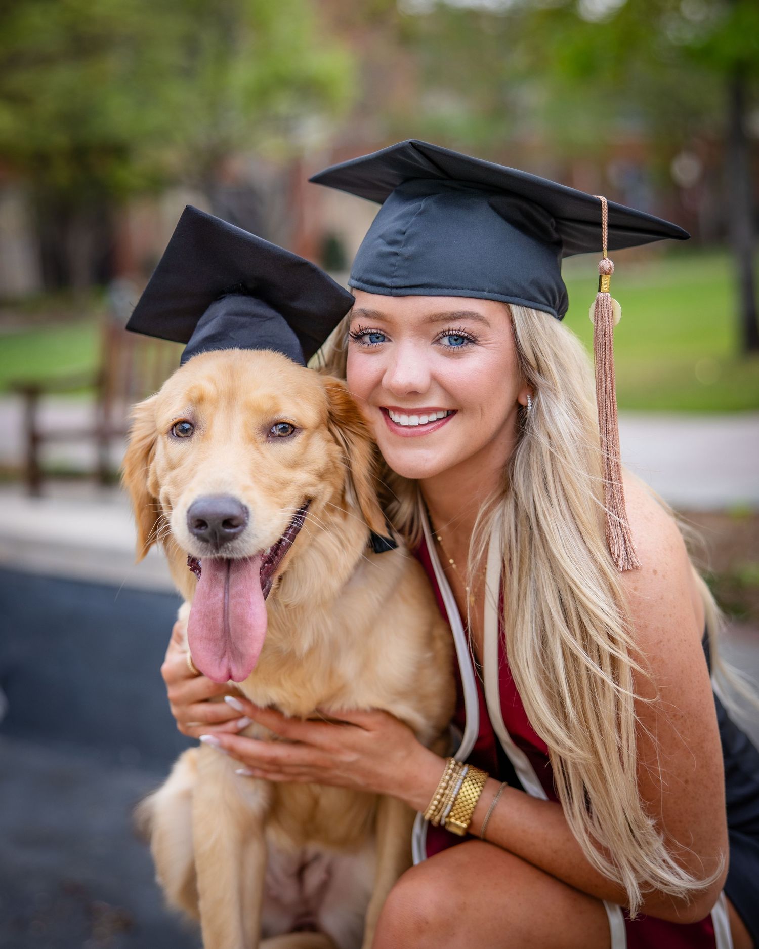 Smiling blonde woman and golden retriever both wearing black graduation caps pose together outdoors for a graduation photo.