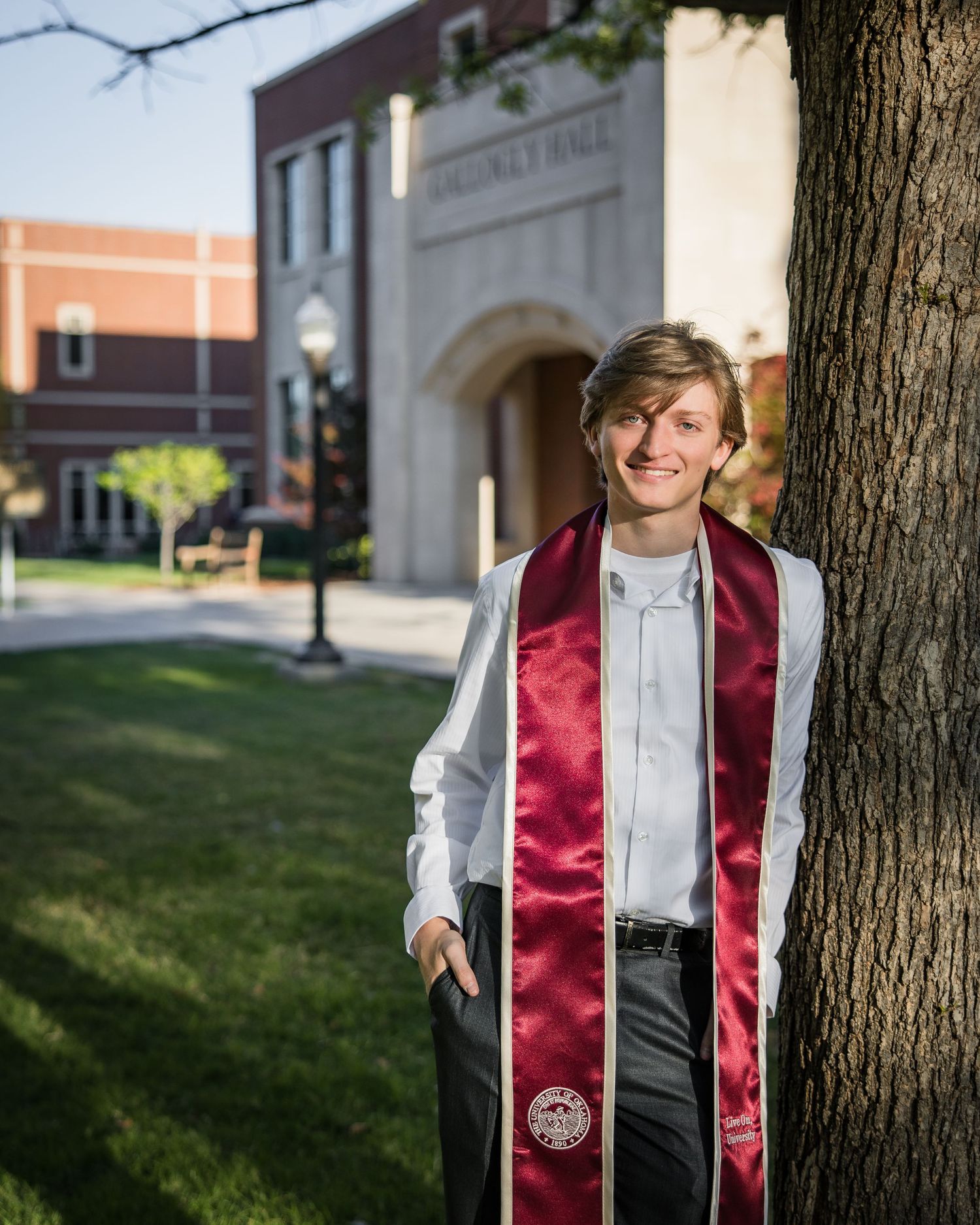 Young man in graduation attire with maroon stole smiling beside a tree on a college campus.