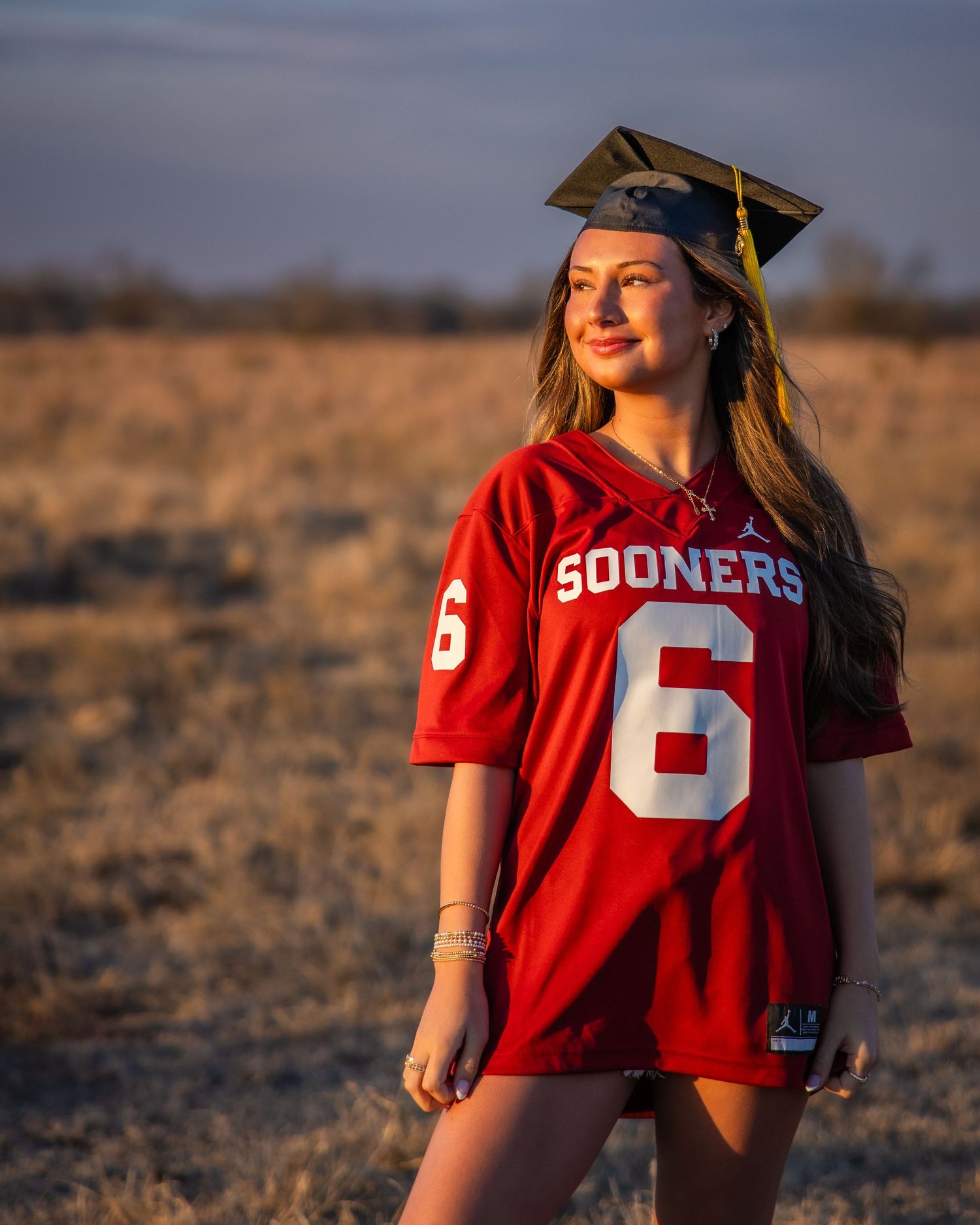 Graduate in red Sooners jersey poses in golden sunlit field during sunset photo session.