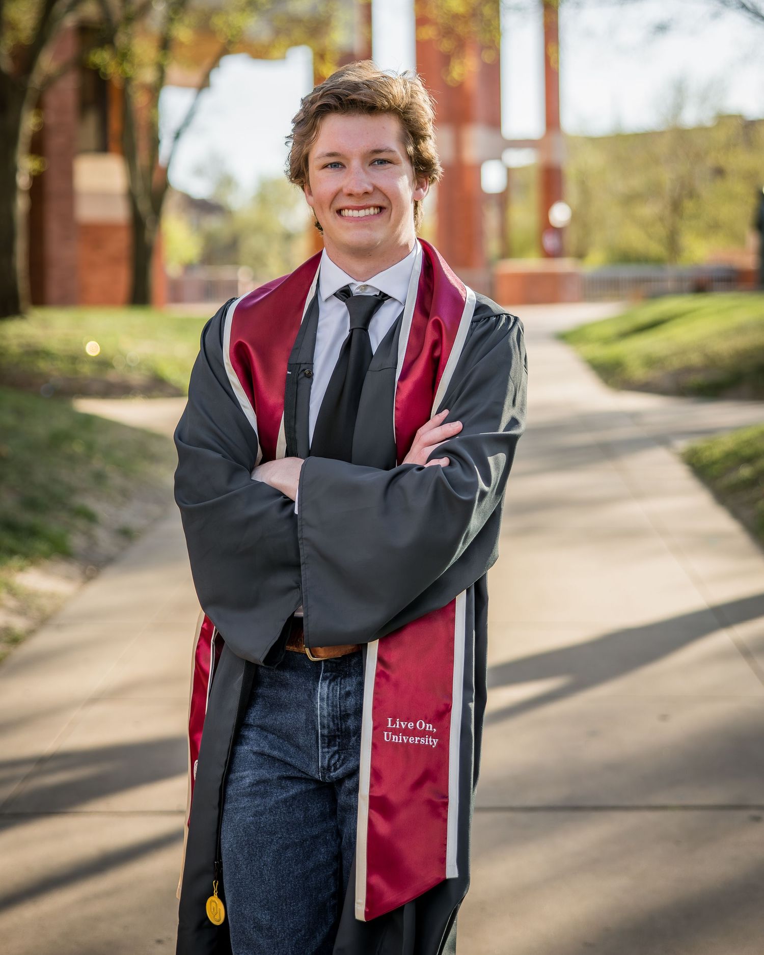 Smiling college graduate in black gown and maroon stole poses with arms crossed on a sunny campus walkway.