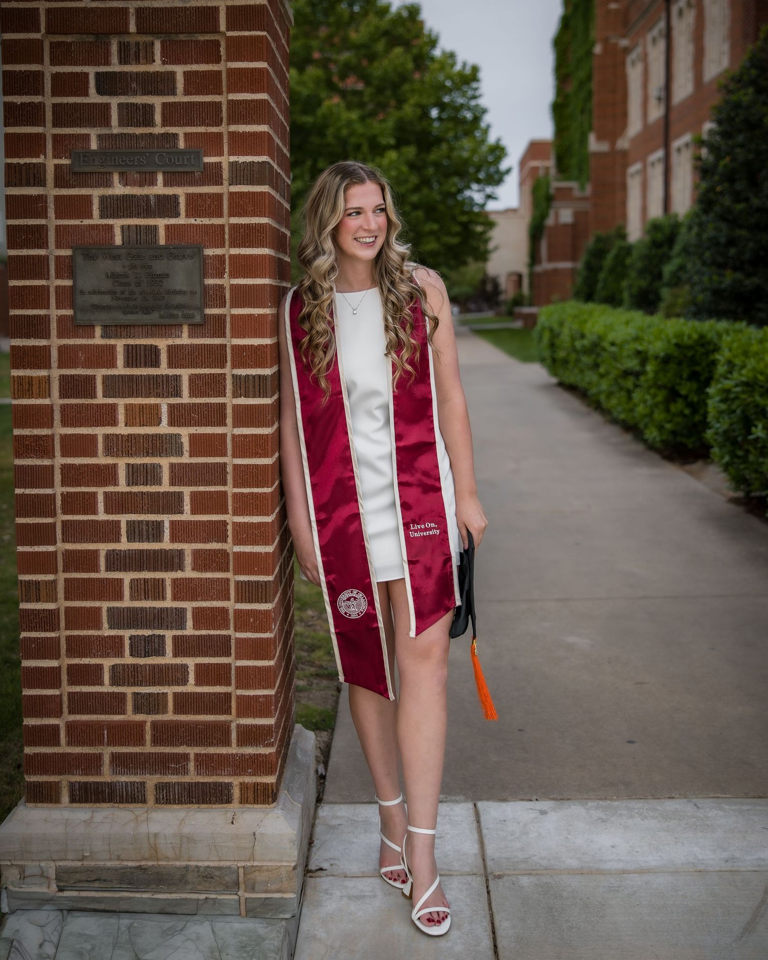 Smiling graduate in white dress and maroon sash poses by brick building on college campus.